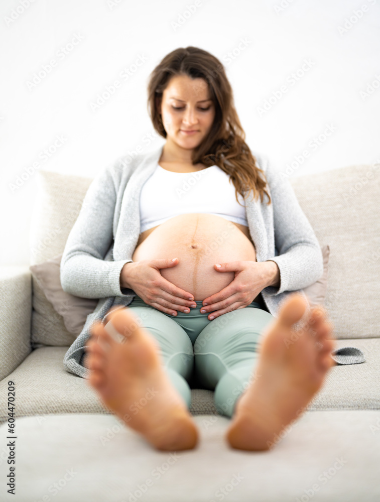 Fototapeta premium Frontal view of smiling woman sitting on a sofa proudly holding her belly in expectation of the baby. Pregnancy third trimester - week 34. Front view. White background.