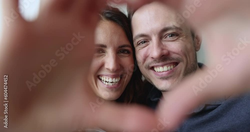 Close up overjoyed, affectionate Hispanic couple looking at camera through joined fingers showing heart symbol. Romantic spouses shoot video, make selfie, celebrate St. Valentine Day. Love, relations