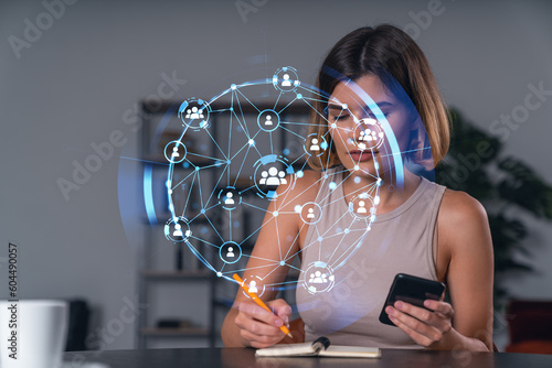 Smiling businesswoman in casual wear holding using smartphone typing at office workplace with notebook. Concept of distant work, business education. Social network icons chain hologram