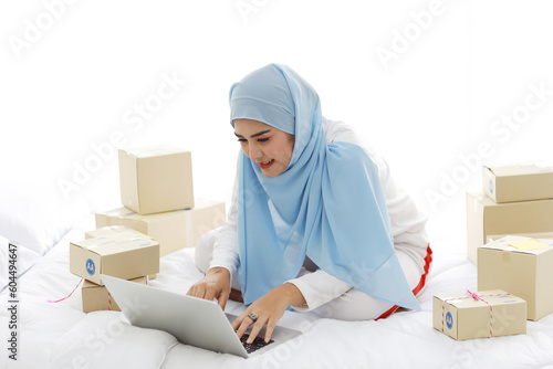 Beautiful and young asian woman in muslim sleepwear with attractive look, sitting on bed with computer and online package box delivery. Startup small business SME freelance girl work with mobile phone © feeling lucky