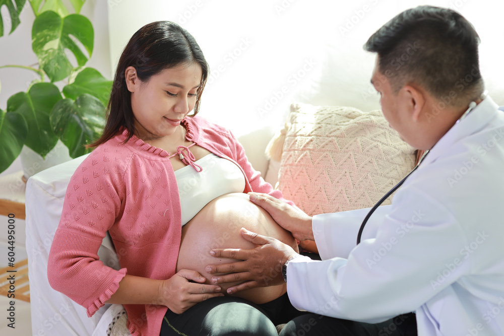 Happy smiling young asian pregnant female and male doctor sitting on sofa in living room at home with private treatment and consultant. Friendly doctor listening to baby’s heartbeat with stethoscope. Happy smiling young asian pregnant female and male doctor sitting on sofa in living room at home with private treatment and consultant. Friendly doctor listening to baby’s heartbeat with stethoscope.