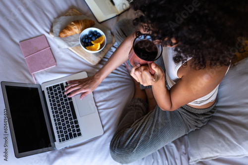 Fotografie High angle view close-up of young unrecognisable woman sitting on bed typing on laptop while having morning coffee