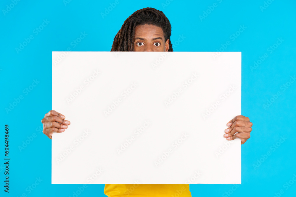 Surprised latin man with dreadlocks hiding behind a blank board Surprised latin man with dreadlocks hiding behind a blank board