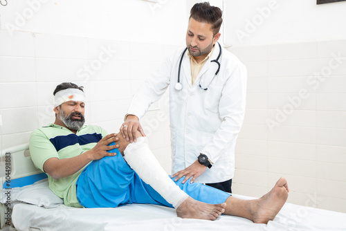 Indian orthopedic doctor examining patient with fractured leg lying on bed at hospital, healthcare concept.