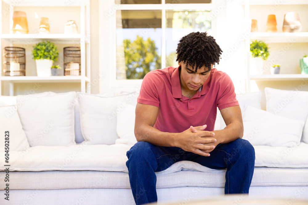 Biracial young man with hands clasped sitting in depression on sofa in living room, copy space
