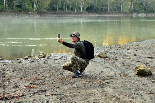 hombre adulto con una mochila negra haciendo fotos y shelfies en un bosque con un lago