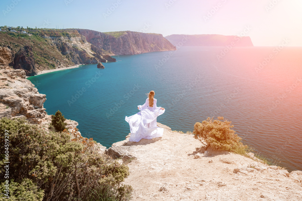 woman sea white dress. Blonde with long hair on a sunny seashore in a white flowing dress, rear view, silk fabric waving in the wind. Against the backdrop of the blue sky and mountains on the seashore woman sea white dress. Blonde with long hair on a sunny seashore in a white flowing dress, rear view, silk fabric waving in the wind. Against the backdrop of the blue sky and mountains on the seashore