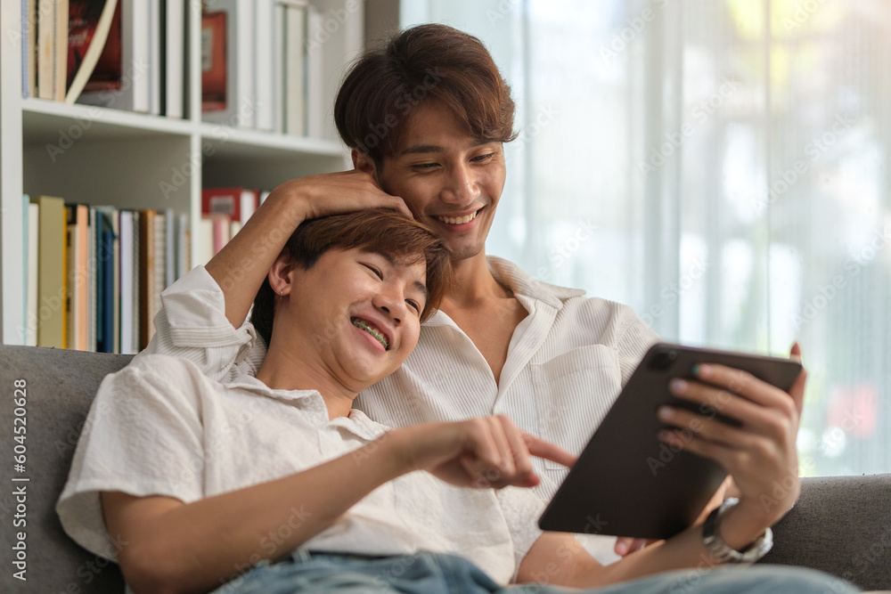 Joyful gay couple sitting on couch shopping online or watching funny videos on digital tablet.