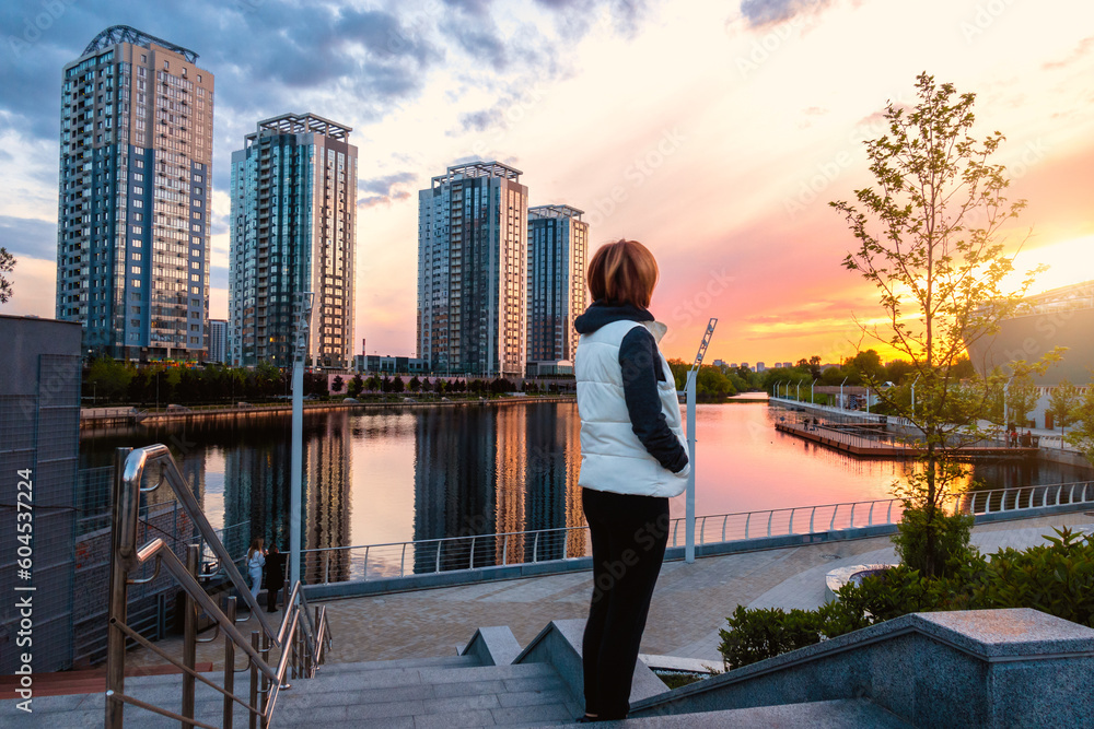 A woman on the embankment of the river in the city looks at the sunset. Kyiv, Rivermall park A woman on the embankment of the river in the city looks at the sunset. Kyiv, Rivermall park