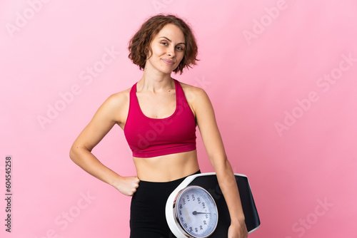 Young English woman isolated on pink background with arms at hip and holding weighing machine
