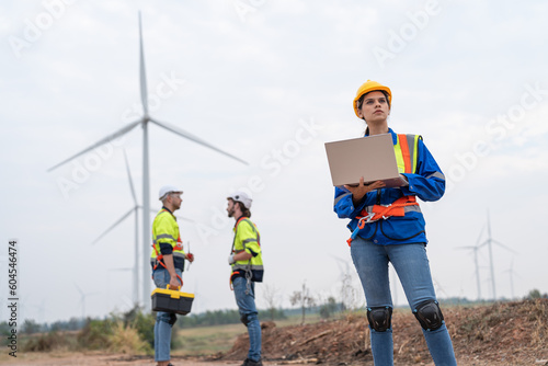 Female engineer in uniform with helmet safety using laptop inspection and maintenance of wind turbines in wind farms to generate renewable energy.