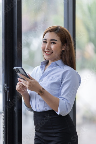 Smiling business Asian woman using her phone in the office. Small business entrepreneur looking at her mobile phone and smiling while communicating with her office colleagues
