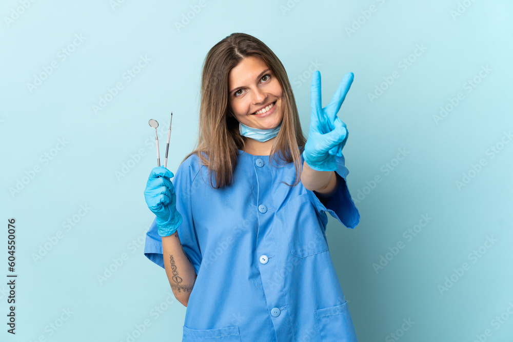 Slovak dentist holding tools isolated on blue background smiling and showing victory sign Slovak dentist holding tools isolated on blue background smiling and showing victory sign