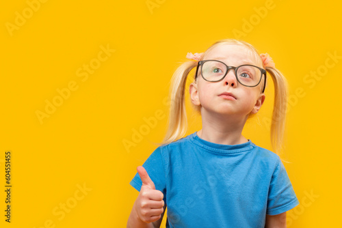 Fototapeta Naklejka Na Ścianę i Meble -  Serious elementary schoolgirl with two ponytails wears blue T-shirt and glasses with thumbs up. Studio portrait of child showing like gesture.