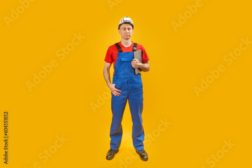Serious male engineer in overalls and hardhat holding on clipboard and looking at camera isolated on yellow background. Copy space.