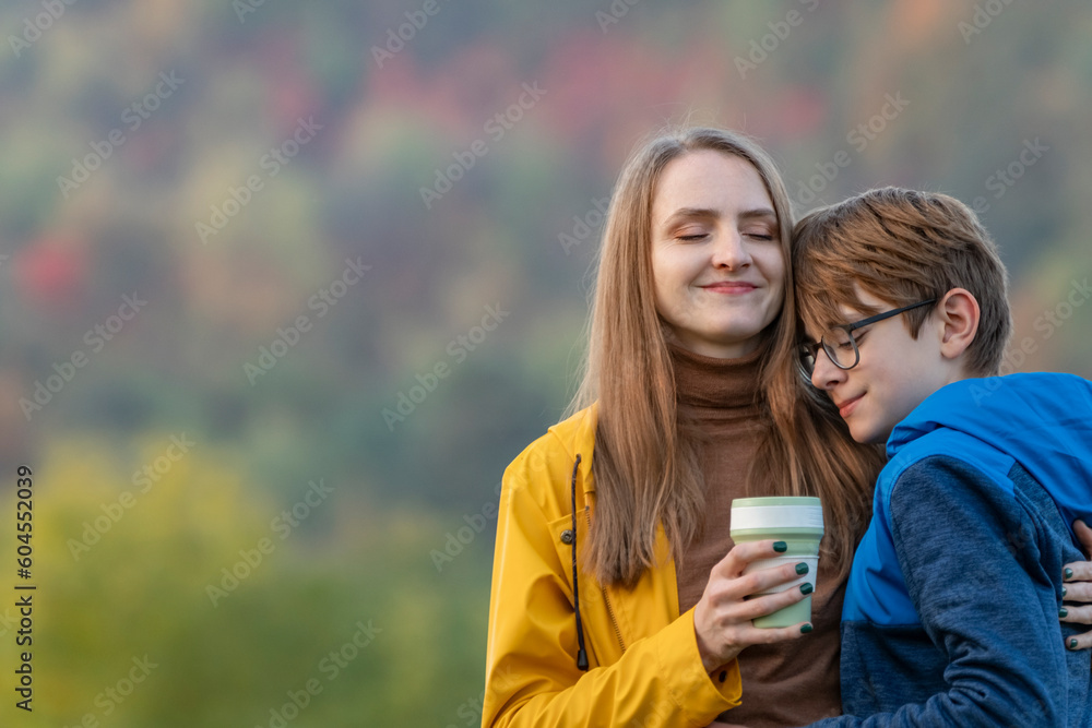 Young mother with cup of hot drink hugs son in nature. Mom and child happily spend autumn day outdoors Young mother with cup of hot drink hugs son in nature. Mom and child happily spend autumn day outdoors
