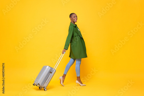 Fashionable skinhead African American woman traveler walking with trolley bag ready to fly against yellow color isolated background studio shot