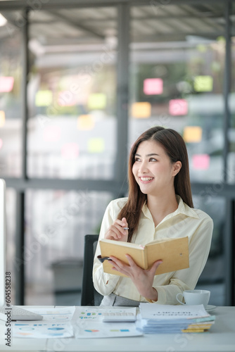 Photo of attractive woman sitting at desk working on computer looking at screen, smiling, wearing yellow shirt holding a book,, typing on keyboard, holding a cup of coffee. 