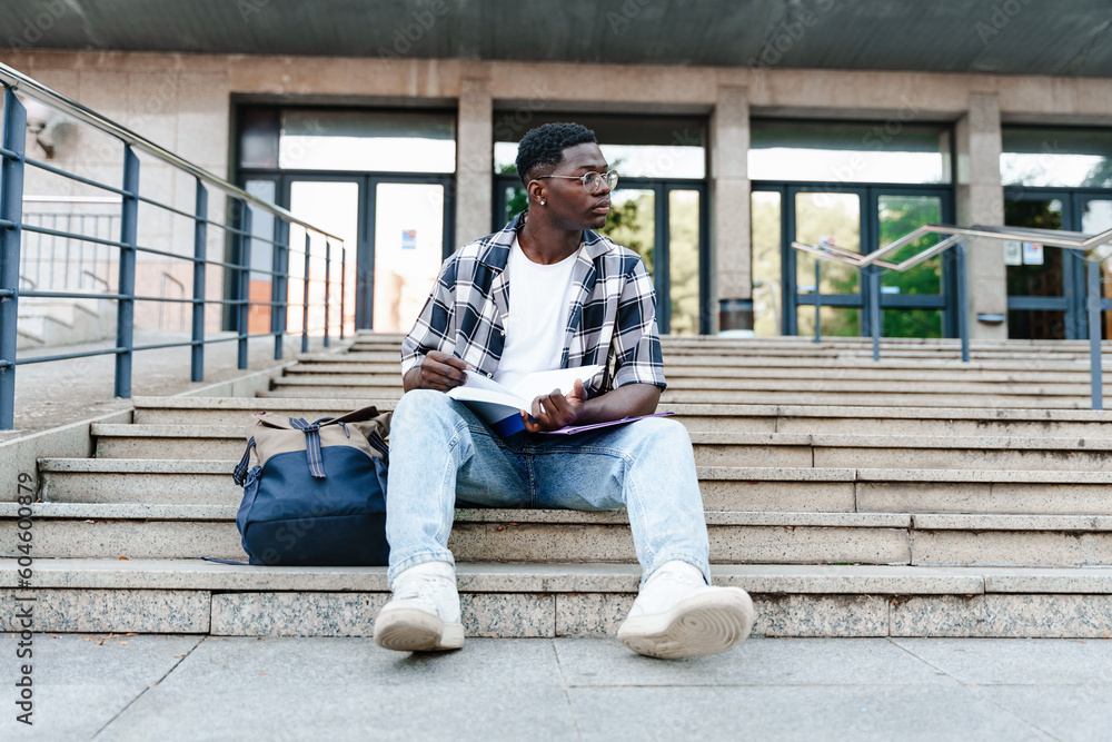 Pensive African student reading book sitting on University Campus staircase Pensive African student reading book sitting on University Campus staircase