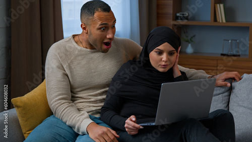 Married ethnic couple husband and wife African american man use laptop with arabian muslim woman at home comfort couch talking buying online freelance shopping win internet discounts rejoice fortune