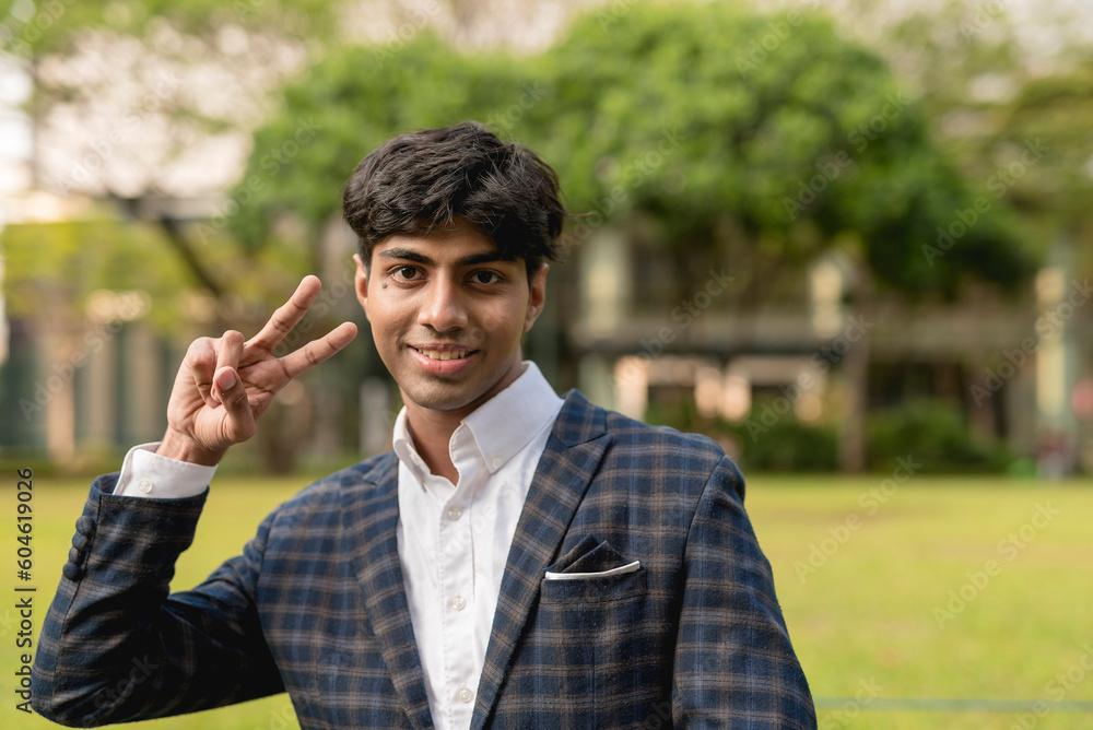 A handsome guy in a white long sleeves and blue checkered suit is smiling at the camera while holding up a peace sign while standing in a greenery area. Trees, house and grass in the background. A handsome guy in a white long sleeves and blue checkered suit is smiling at the camera while holding up a peace sign while standing in a greenery area. Trees, house and grass in the background.