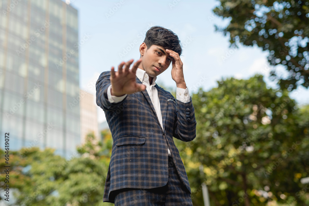 A stylish guy in a blue checkered suit in a serious face while looking at the camera, his left hand on his forehead and the other is reaching out. He's standing outside, in the city. A stylish guy in a blue checkered suit in a serious face while looking at the camera, his left hand on his forehead and the other is reaching out. He's standing outside, in the city.