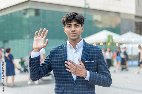 A stylish guy in a blue checkered suit in an oath taking pose with a serious face while standing outside, in the city. Building, people and tents are shown in the background.