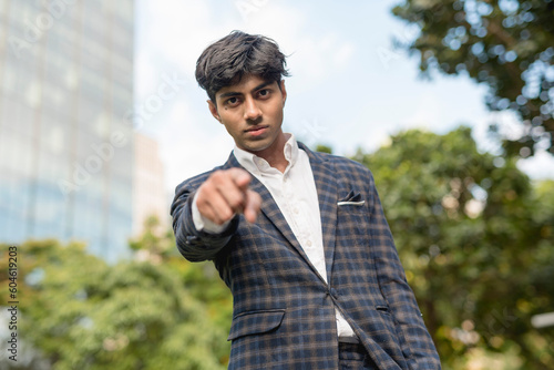A stylish guy in a blue checkered suit is looking and pointing at the camera with a serious face while standing outside. A building, sky and trees in the background.