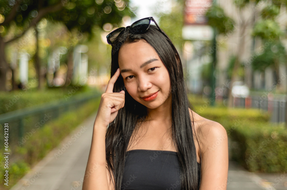 A pretty lady standing in the park wearing a black strapless tank top and sunglasses in a thinking pose smiling while standing outside, in the park. A trees, fence and bushes in the background. A pretty lady standing in the park wearing a black strapless tank top and sunglasses in a thinking pose smiling while standing outside, in the park. A trees, fence and bushes in the background.