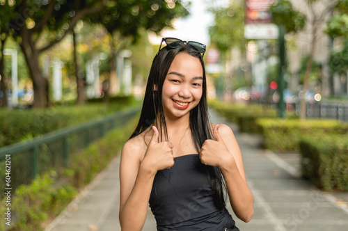 A pretty young lady standing in the park wearing a black strapless tank top and sunglasses, smiling while holding up a two thumbs up hand gesture. Trees,bushes and fence in the background.