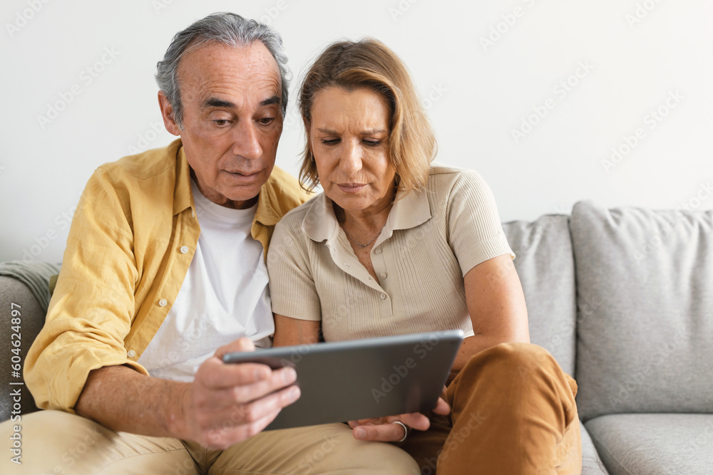 Puzzled senior spouses using digital tablet having hard time learning or browsing internet, sitting on sofa, copy space