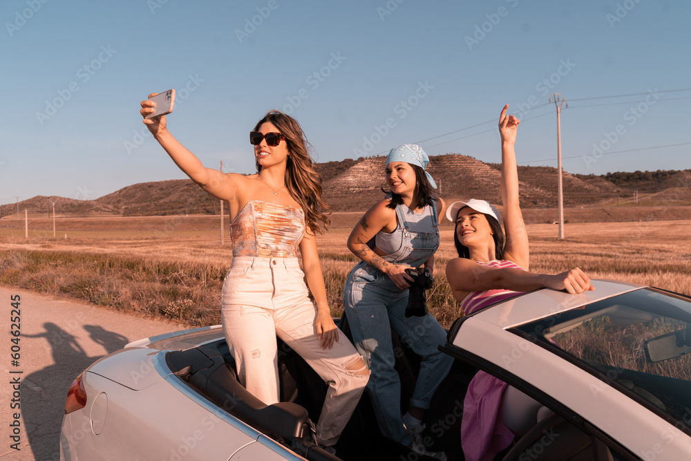 The pretty girl with wavy hair taking a selfie with her friend dressed in a blue jumpsuit and scarf holding a camera and her other friend dressed in pink clothes, while traveling on summer vacation.