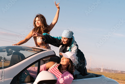 The three young friends are on a summer trip.The girl in pink clothes is driving the convertible and the girl with the scarf is pointing to the next destination.