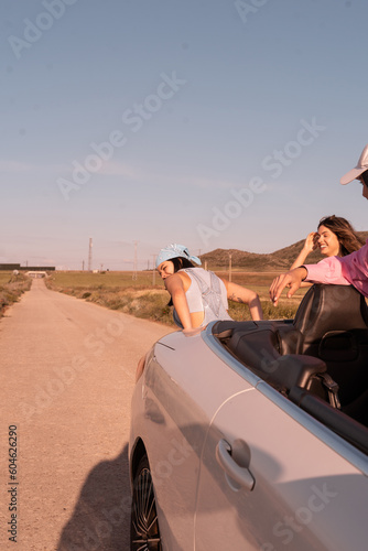 The girl with the blue scarf on her head tries to start the broken down white car and her friend cheers them on from inside the car ,they are on a road on their summer vacation.