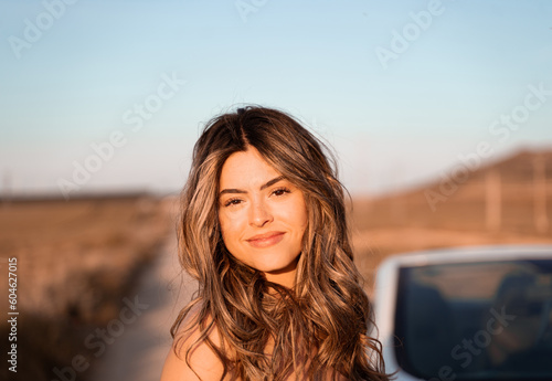 The young woman with white skin and wavy hair smiles at the camera, she is in the countryside on a summer afternoon.