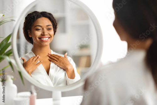 Black smiling woman looking in mirror and touching decollete zone photo