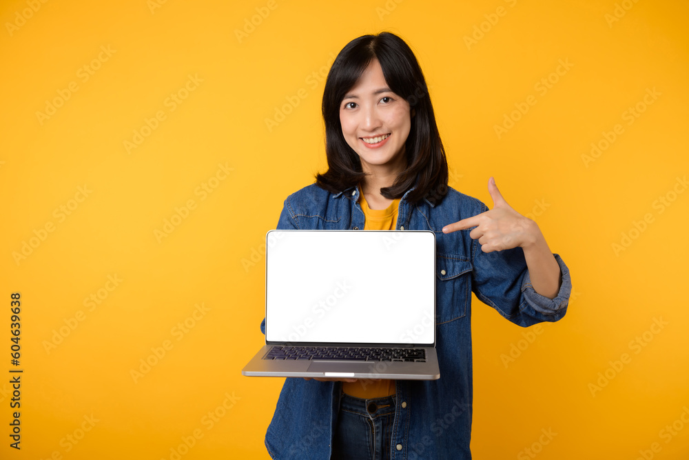 portrait young happy woman wearing yellow t-shirt and denim shirt holding laptop and point finger to screen isolated on yellow studio background. business technology application communication concept. portrait young happy woman wearing yellow t-shirt and denim shirt holding laptop and point finger to screen isolated on yellow studio background. business technology application communication concept.