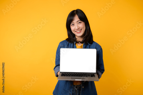 portrait young happy woman wearing yellow t-shirt and denim shirt holding laptop and point finger to screen isolated on yellow studio background. business technology application communication concept. © Jirawatfoto