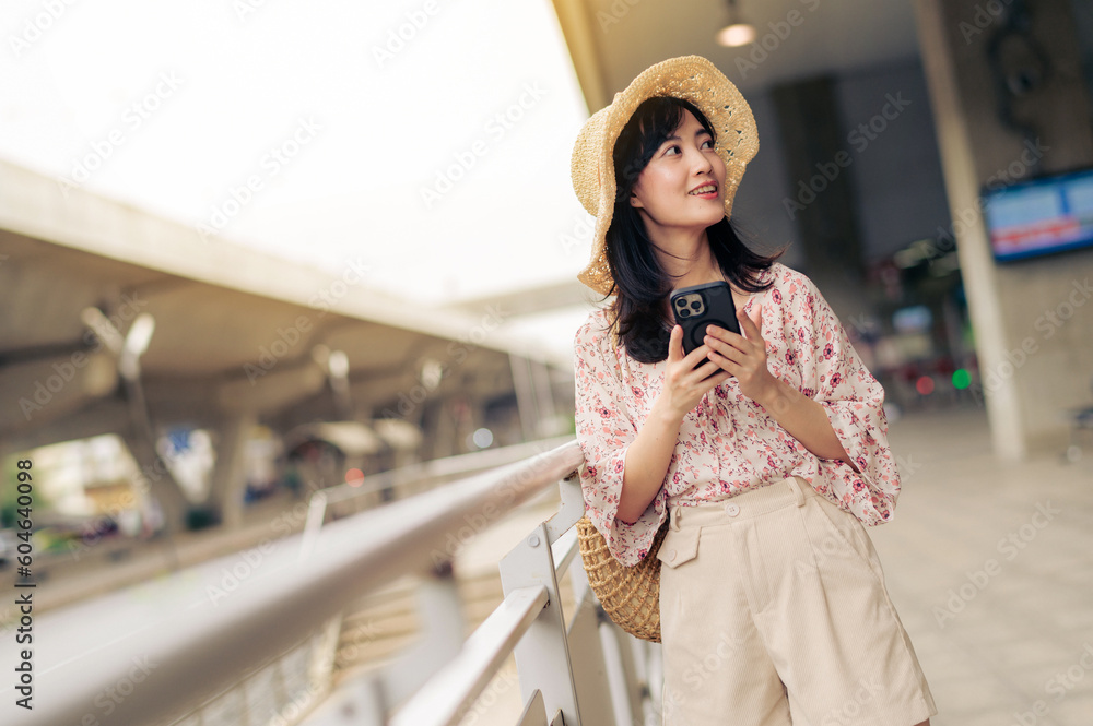 Asian young woman traveler with weaving basket using a mobile phone beside railway train station in Bangkok. Journey trip lifestyle, world travel explorer or Asia summer tourism concept.