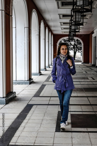 woman walks in arched gallery. Pretty woman in blue coat and jeans walking in arched gallery of building
