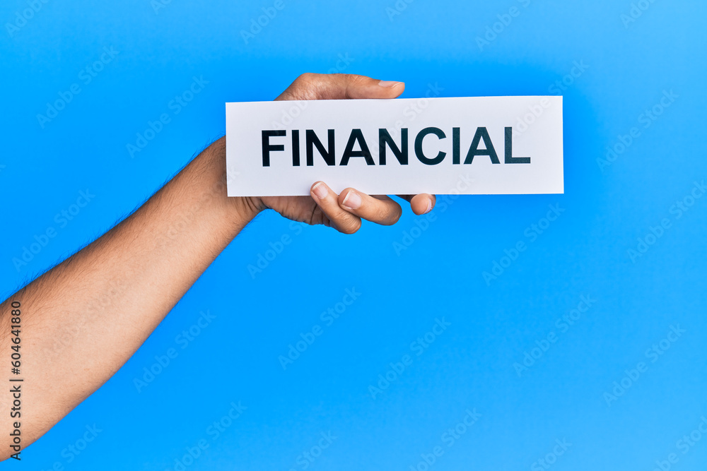 Hand of caucasian man holding paper with financial word over isolated blue background Hand of caucasian man holding paper with financial word over isolated blue background