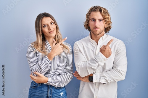 Young couple standing over blue background pointing with hand finger to the side showing advertisement, serious and calm face