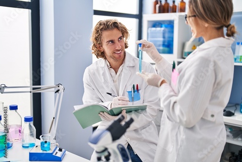 Man and woman wearing scientist uniform holding test tube writing on notebook at laboratory