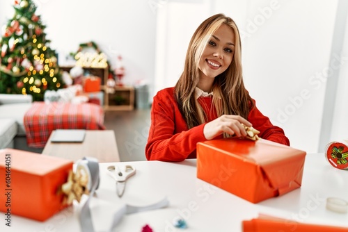 Young caucasian woman preparing gift sitting by christmas tree at home