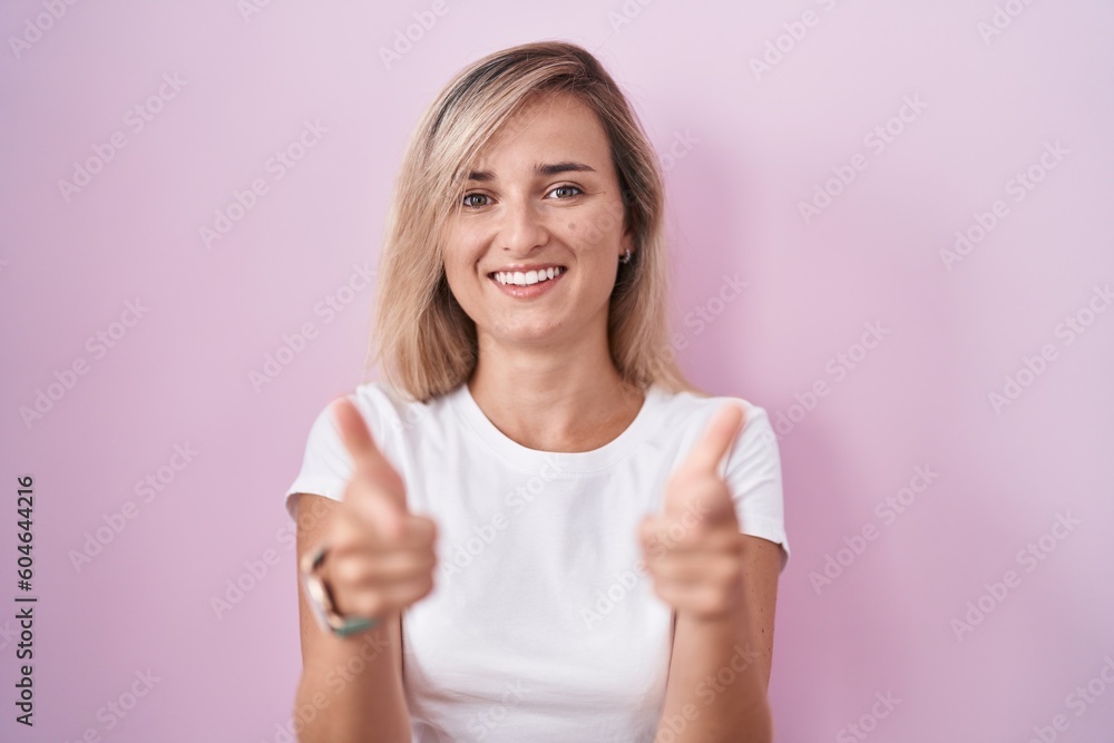 Young blonde woman standing over pink background pointing fingers to camera with happy and funny face. good energy and vibes. Young blonde woman standing over pink background pointing fingers to camera with happy and funny face. good energy and vibes.