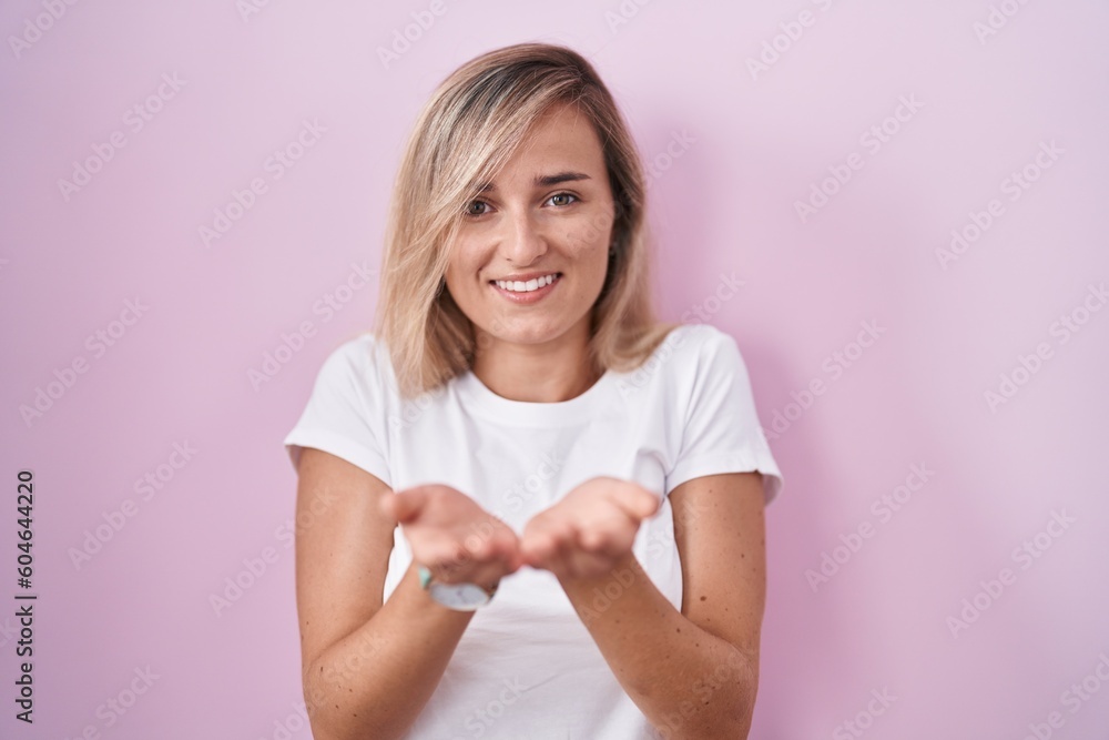 Young blonde woman standing over pink background smiling with hands palms together receiving or giving gesture. hold and protection Young blonde woman standing over pink background smiling with hands palms together receiving or giving gesture. hold and protection