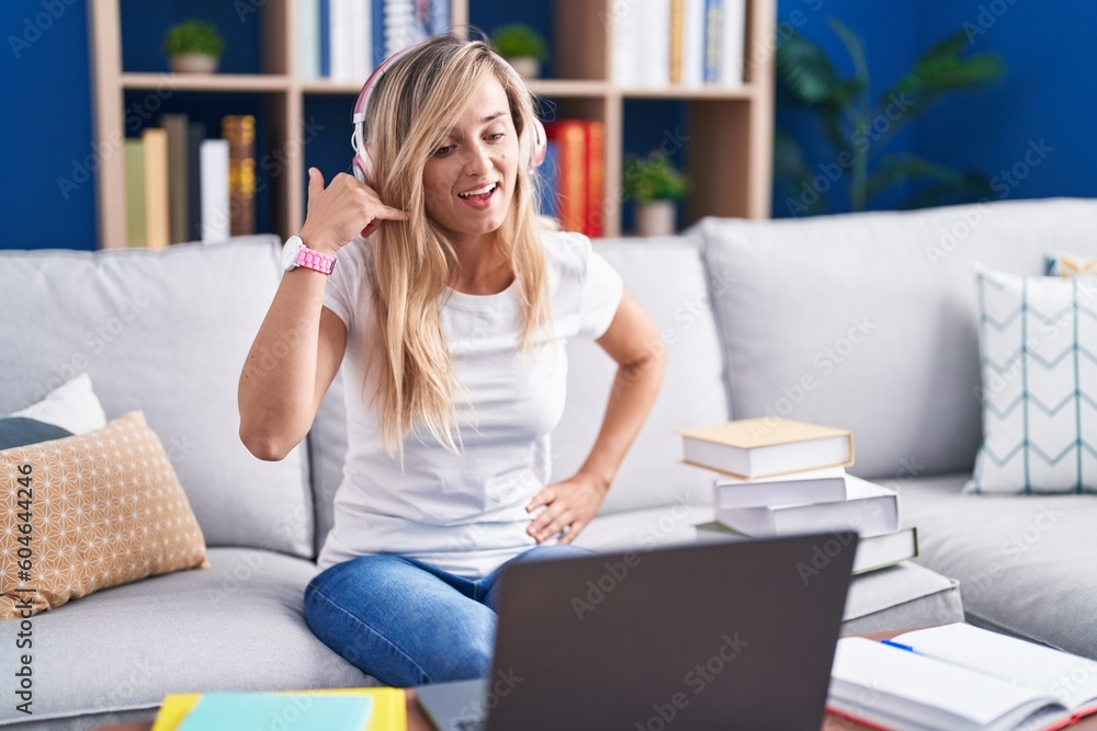 Young blonde woman studying using computer laptop at home smiling doing phone gesture with hand and fingers like talking on the telephone. communicating concepts. Young blonde woman studying using computer laptop at home smiling doing phone gesture with hand and fingers like talking on the telephone. communicating concepts.