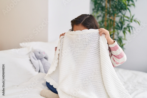 Adorable hispanic girl sitting on bed covering face with blanket at bedroom