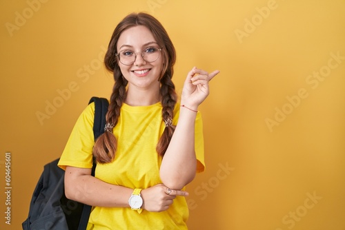 Young caucasian woman wearing student backpack over yellow background with a big smile on face  pointing with hand finger to the side looking at the camera.
