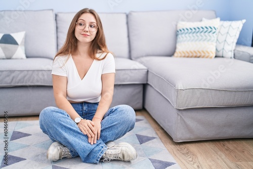 Young caucasian woman sitting on the floor at the living room smiling looking to the side and staring away thinking.